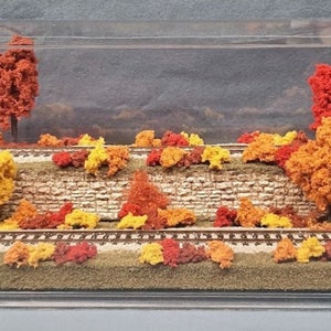 May include: A miniature model railroad scene with a stone wall and a track surrounded by fall foliage. The foliage is made of red, orange, and yellow felt.