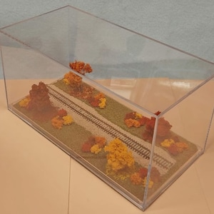 May include: A miniature model railroad scene in a clear acrylic display case. The scene features a pair of train tracks running through a grassy area with fall foliage in shades of orange, yellow, and brown.