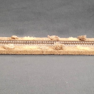 May include: A model railroad track with brown and tan grass-like material on either side of the track. The track is made of metal and has two rails.