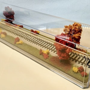 May include: A clear acrylic display case with a miniature model railroad scene inside. The scene features a track with fall foliage in shades of red, orange, and yellow.