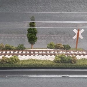 May include: A miniature model railroad scene in a clear acrylic display case. The scene features a small train track with a crossing sign, trees, and a grassy area.