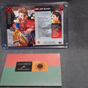 May include: A clear plastic case with a trading card of Jeff Gordon, a NASCAR driver, in a red and yellow race car. The card is from the 2002 season and is autographed by Jeff Gordon. The card is on a black background with white text. The case is on a gray surface.
