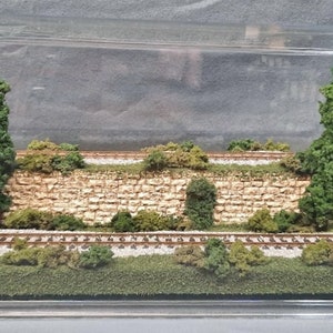 May include: A miniature model railroad scene with a stone wall, green trees, and a track with a grassy area. The scene is displayed in a clear plastic case.