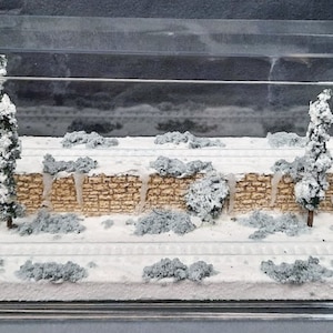 May include: A miniature winter scene with a stone wall, snow-covered trees, and a snowy ground. The scene is displayed in a clear plastic case.