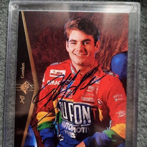May include: A signed Jeff Gordon NASCAR trading card featuring a close-up of the driver in his racing uniform. The card has a gold border and a black background with white text. The driver is wearing a red, white, and blue racing suit with the words "U-FON" and "AUTOMOTIVE PAINTS" on the front. The card is signed in black ink.
