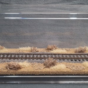 May include: A clear acrylic display case with a miniature model railroad track and brown and tan foliage inside.