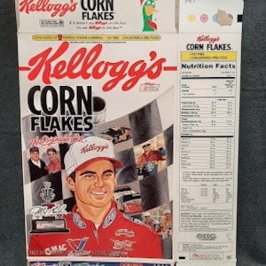 Könnte beinhalten: Eine Kellogg's Corn Flakes-Müslipackung mit dem NASCAR-Fahrer Jeff Gordon. Die Packung ist rot, weiß und blau mit einem Bild von Gordon in seinem Rennwagen. Die Packung enthält auch das Kellogg's-Logo und die Worte "Corn Flakes".