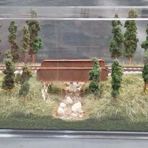 May include: A miniature model railroad scene with a wooden bridge, trees, and a grassy area. The scene is displayed in a clear acrylic case.