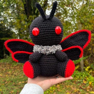 以下が含まれることがあります： 赤いアクセントと大きな赤い目をつけた黒い編み物の蝶。蝶は首に灰色の編み物のスカーフをしています。蝶は人の手に持たれています。*PATTERN*
