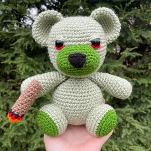 May include: A crocheted green and gray teddy bear with red eyes and a brown and orange stick in its hand.