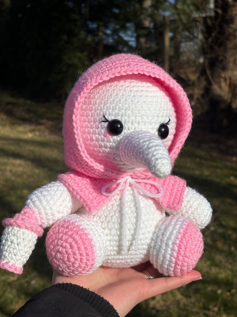 Pu&ograve; includere: Un giocattolo di peluche di uccello all'uncinetto rosa e bianco con un mantello con cappuccio rosa. L'uccello ha un lungo becco bianco e occhi neri.