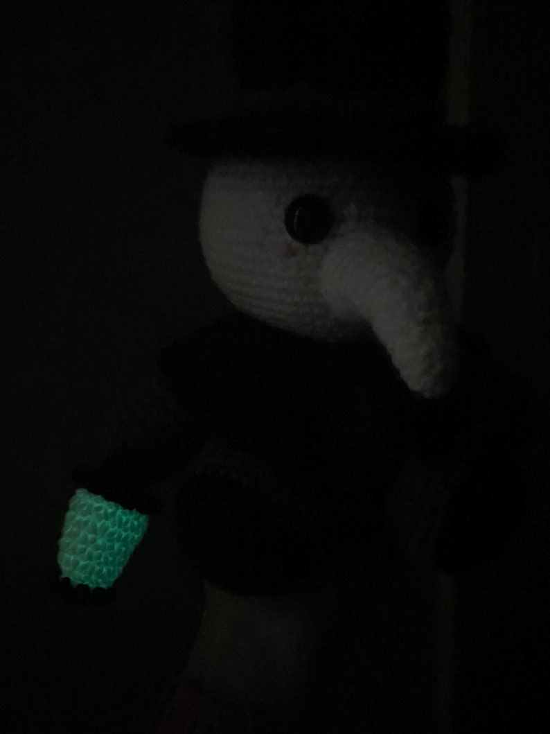 Pu&ograve; includere: Un giocattolo di peluche all'uncinetto con un cappello a cilindro nero, un viso bianco e un lungo naso nero. Il giocattolo tiene un oggetto verde luminoso nella sua mano destra.