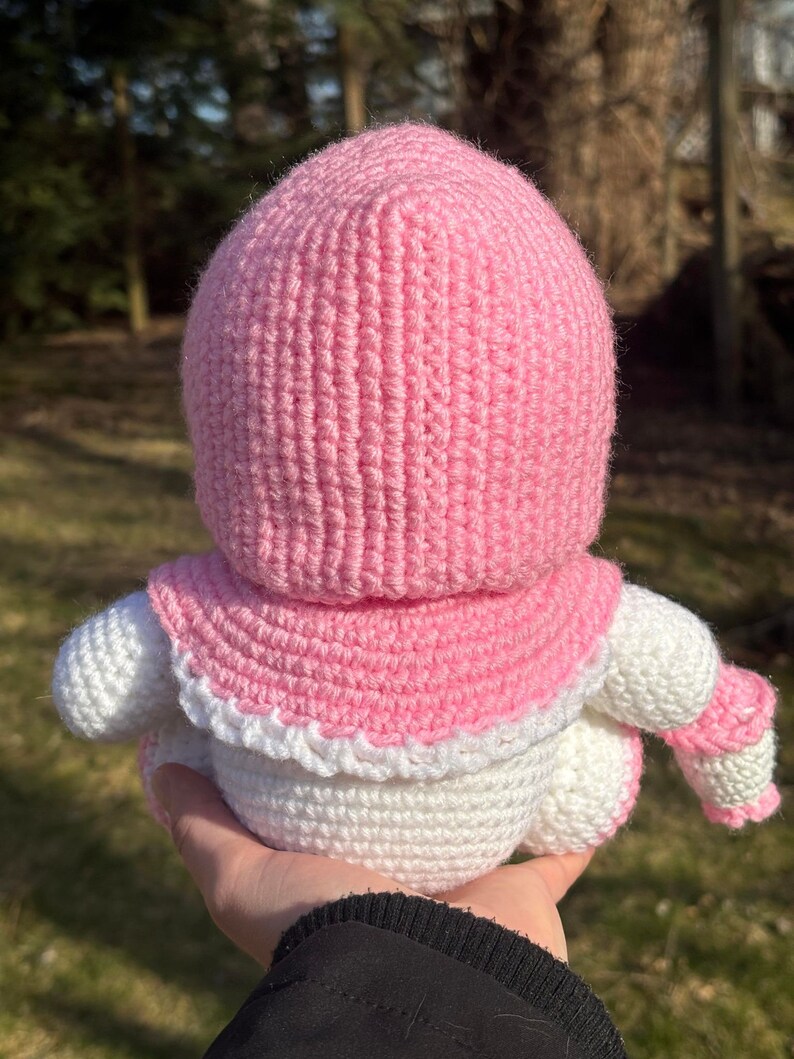 Pu&ograve; includere: Una bambola all'uncinetto con un cappello con cappuccio rosa e un corpo bianco. La bambola indossa una sciarpa all'uncinetto bianca.