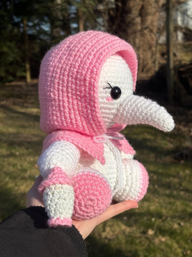Pu&ograve; includere: Un peluche lavorato a uncinetto con un cappello con cappuccio rosa, corpo bianco e dettagli rosa. L'animale ha un naso lungo e bianco e occhi neri.