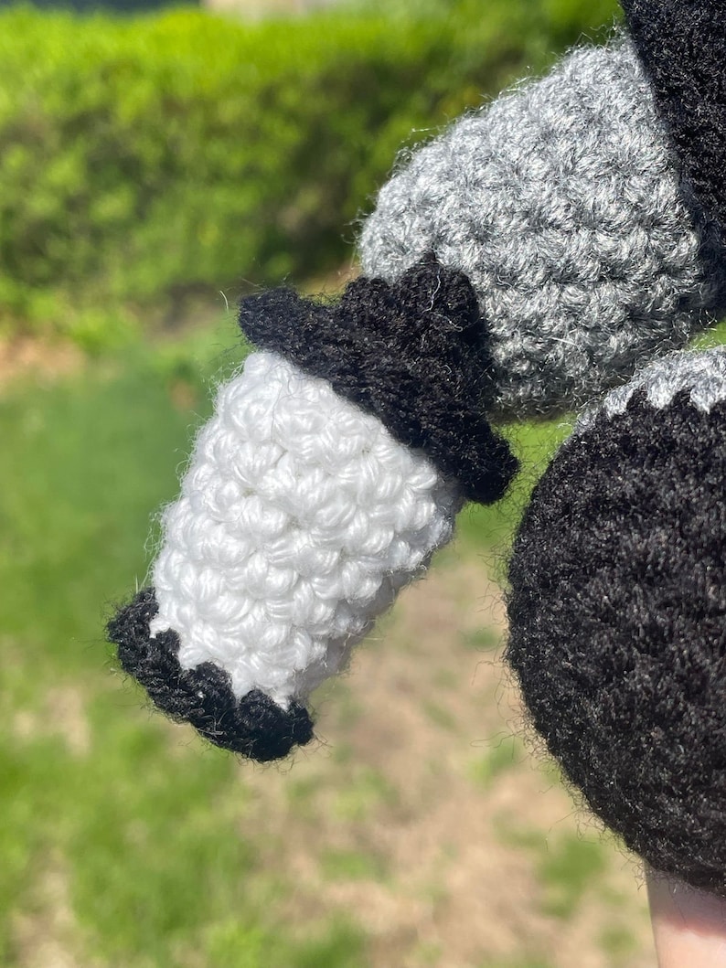 Pu&ograve; includere: Primo piano di un giocattolo amigurumi all'uncinetto con filato nero, bianco e grigio. Il giocattolo ha un corpo bianco, una testa nera e un braccio grigio.