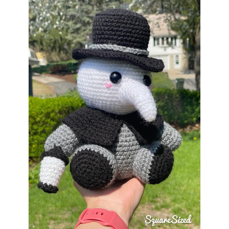 Pu&ograve; includere: Un giocattolo di peluche all'uncinetto a forma di personaggio che indossa un cappello a cilindro nero, un cappotto nero e grigio e un lungo naso bianco. Il giocattolo ha occhi neri e guance rosa.