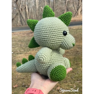 May include: A green crocheted dinosaur toy with black eyes. The dinosaur is sitting on a hand.
