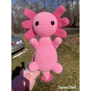 May include: A handmade, pink crochet axolotl plushie with black safety eyes. The axolotl has six frilly, petal-like gills on each side of its head, and small arms and legs. The plushie is approximately 10 inches tall.
