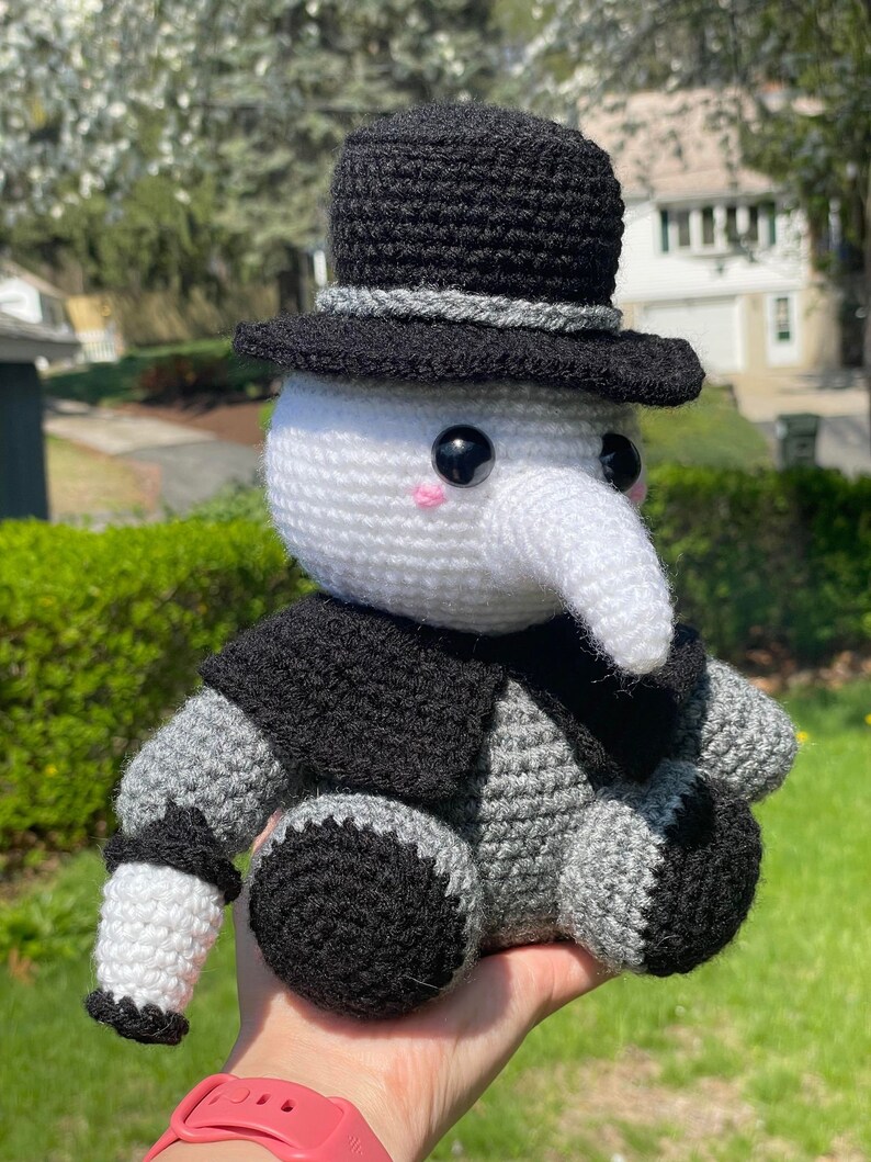 Pu&ograve; includere: Un giocattolo di peluche all'uncinetto a forma di persona che indossa un cappello a cilindro nero e una maschera bianca. Il giocattolo ha un naso lungo e bianco e occhi neri. Il giocattolo &egrave; seduto nella mano di una persona.