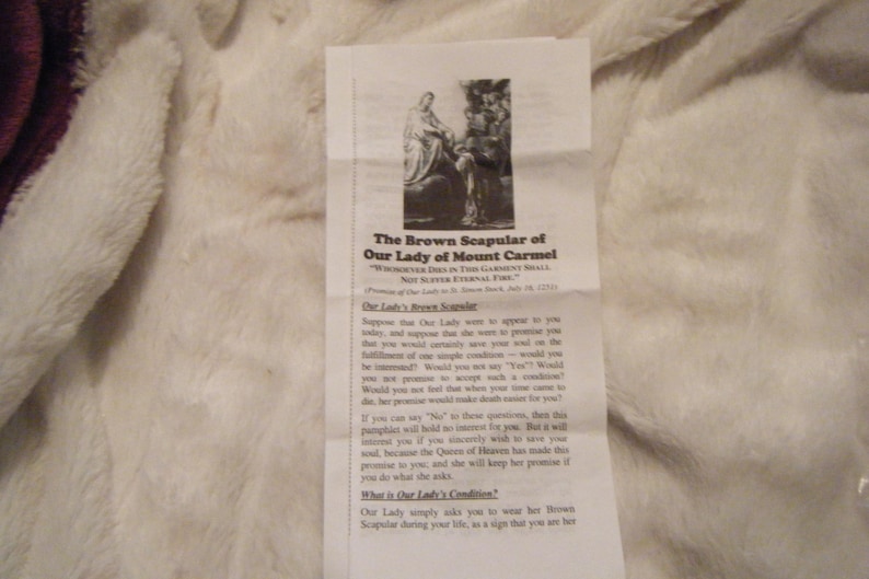 May include: A printed pamphlet titled "The Brown Scapular of Our Lady of Mount Carmel" with an image of a religious figure. The text is in black font on a white background. The pamphlet is lying on a white, fluffy surface.