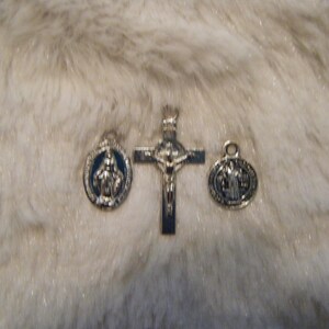 Puede incluir: Tres amuletos religiosos de plata: un crucifijo con una figura de Jesucristo, un amuleto redondo con una representaci&oacute;n de la Virgen Mar&iacute;a y un amuleto redondo con una representaci&oacute;n de San Crist&oacute;bal.