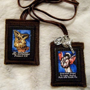 May include: Two brown fabric prayer cards with images of angels and text. The first card features an image of the Archangel Michael with the text "St. Michael The Archangel Protect Us!" The second card features an image of a guardian angel with the text "Guardian Angel To Light, Guard Rule and Guide Me".
