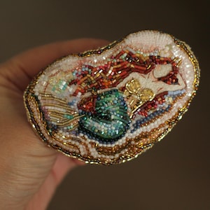 May include: An oval-shaped brooch featuring intricate beadwork embroidery of a mermaid with flowing red hair and a shimmering green tail. The design is accented with gold, white, and iridescent beads, creating a textured, multi-dimensional surface.