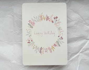 May include: A white card with a floral wreath design in shades of pink, purple, and green. The text "happy birthday" is written in the center of the wreath.