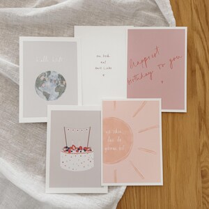 Puede incluir: Un conjunto de tarjetas de felicitación con varios diseños y texto. Una tarjeta presenta un mapa del mundo, otra dice "Happiest Birthday to you", y otras tienen ilustraciones de pastel y sol. Las tarjetas son de colores pastel suaves.