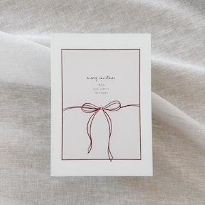 Könnte beinhalten: Weiße Weihnachtskarte mit burgunderrotem Rand und Schleifenillustration. Der Text "merry christmas" steht über "from our family to yours". Die Karte ist rechteckig und hat ein minimalistisches Design.