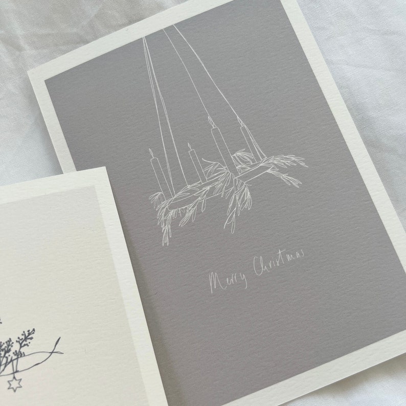 May include: A gray card with a white line drawing of a candle chandelier with greenery hanging from it. The text "Merry Christmas" is written in white below the drawing.