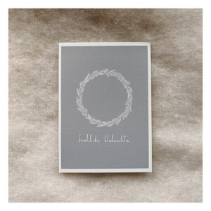 Peut inclure: Carte de vœux avec un fond gris et un bord blanc. La carte présente un motif de couronne circulaire et les mots "fröhliche Weihnachten" en écriture blanche. La carte est sur une surface texturée de tons neutres.