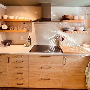May include: A modern kitchen with light wood cabinets, open shelving, and a stainless steel range hood. The shelves display white dishes, bowls, and glassware. A black cooktop is set into the countertop. The kitchen is well-lit with natural light.