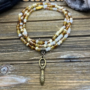 May include: A layered beaded necklace with a bronze pendant. The necklace features three strands of small, colorful beads in shades of brown, cream, and gold. The pendant is a stylized female figure with a spiral design. The necklace is displayed on a wooden surface.