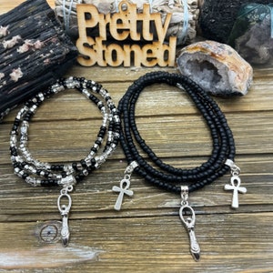 May include: Two beaded bracelets with silver charms. One bracelet features black and clear beads, the other is black. Charms include an Ankh cross and a stylized figure. The wooden sign in the background reads "Pretty Stoned."