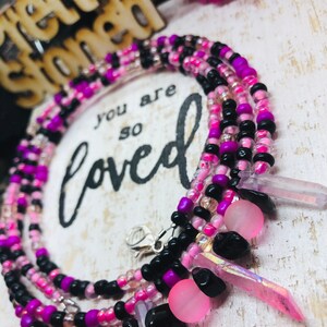 May include: A multi-strand beaded necklace in shades of pink, purple, and black. The necklace features small beads, crystal accents, and a silver clasp. The background includes the text "you are so loved".