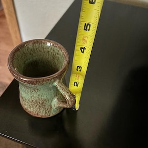 May include: A small, handmade ceramic mug with a rounded body and a handle. The mug is approximately 3 inches tall. It features a speckled green glaze with brown accents and a brown rim.