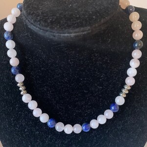 Puede incluir: Un collar hecho con cuentas de piedras preciosas rosas y azules. El collar tiene detalles plateados.