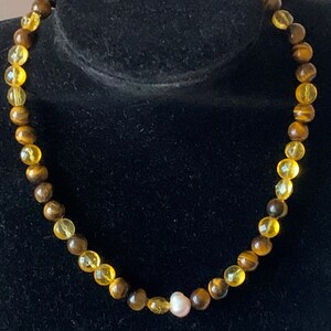 Puede incluir: Un collar hecho de cuentas de ojo de tigre marrón y dorado con una sola cuenta de perla blanca.