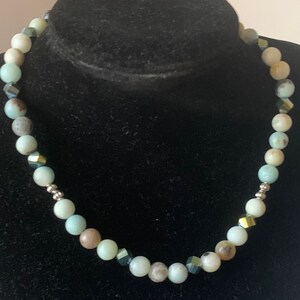 Puede incluir: Un collar de cuentas azul claro y marrón con detalles dorados y negros. El collar está hecho de piedras naturales y mide aproximadamente 45 cm de largo.