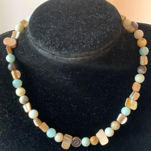 Puede incluir: Un collar de cuentas multicolores con una variedad de formas y tamaños. Las cuentas son en tonos de marrón, beige, blanco y azul claro. El collar está en un maniquí negro.