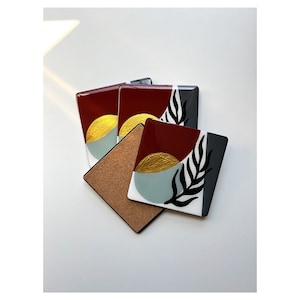 May include: Set of four coasters with abstract geometric designs. The coasters feature a combination of red, gold, black, white, and blue colors. The coasters are made of glass and have a cork backing.