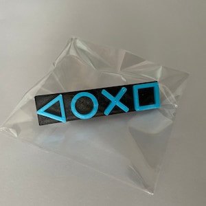May include: A black plastic bar with four blue symbols: a triangle, a circle, an X, and a square. The symbols are commonly used on video game controllers.