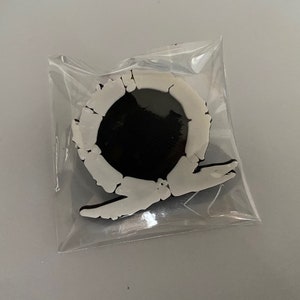 Puede incluir: Un pin de plástico blanco con un círculo negro en el centro y un diseño estilizado alrededor del círculo. El pin está en una bolsa de plástico transparente.