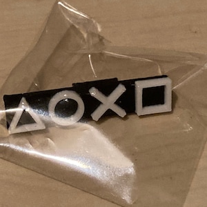 Può includere: Un portachiavi in plastica nera con simboli bianchi del controller PlayStation: un triangolo, un cerchio, una croce e un quadrato.