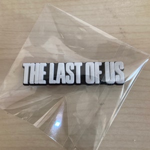 Könnte beinhalten: Ein schwarzer und weißer Plastik-Pin mit dem Text "The Last of Us" in weißen Buchstaben.