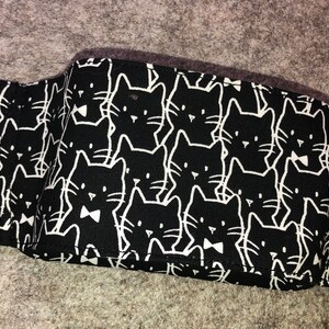 May include: Black fabric face mask with a white repeating pattern of cartoon cats. The cats have simple features and some are wearing bow ties.