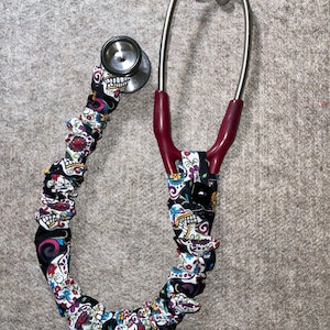 May include: A medical stethoscope with a decorative cover featuring a colorful sugar skull pattern on a black background. The stethoscope has a silver chest piece and earpieces, and a burgundy tube.