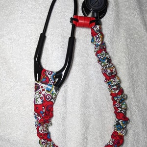 Peut inclure: Un stéthoscope noir avec un revêtement en tissu à motifs rouge et blanc. Le revêtement présente un motif de crâne et de fleurs.