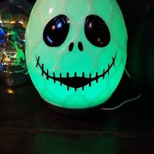 May include: A green and black Halloween-themed essential oil diffuser shaped like a skull with a toothy grin. The diffuser is lit up with a green glow.